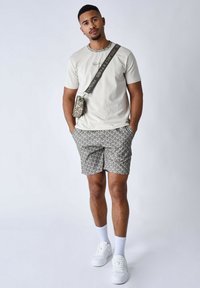 Camiseta de color beige claro con cuello redondo. Pantalones cortos grises con un diseño abstracto. Zapatillas blancas y calcetines blancos hasta el tobillo. Bolso cruzado.