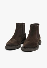 Para ciemnych brązowych zamszowych botków Chelsea z detalami brogu i elastycznymi bocznymi panelami na białym tle.