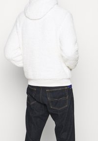 Giacca bianca con cappuccio e tessuto soffice, dal design corto, abbinata a jeans di denim scuro con cuciture evidenti e tasche posteriori.