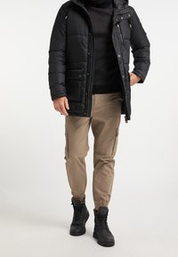 Svart puffjacka, ribbad svart tröja, beige cargobyxor med sidofickor och svarta snörskor. Funktionell utomhusklädsel.