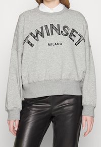 Sweatshirt gris avec le texte "TWINSET MILANO" porté sur une chemise blanche à col, associé à un pantalon en cuir noir.
