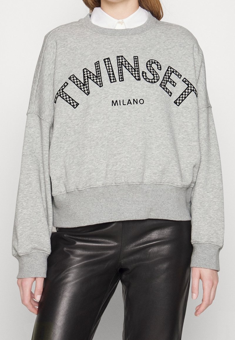 Sweatshirt gris avec le texte "TWINSET MILANO" porté sur une chemise blanche à col, associé à un pantalon en cuir noir.