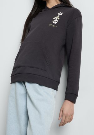 Jeune personne portant un sweat à capuche noir avec des motifs floraux et de cœur sur la poitrine, associée à un jean large bleu clair, debout contre un mur blanc.