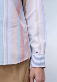 Camicia a righe in colori pastello (blu, rosa, beige) con maniche lunghe. Presenta un polsino a righe in contrasto e un piccolo emblema ricamato.