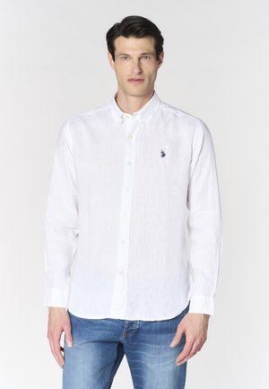 Uomo che indossa una camicia bianca con bottoni e un piccolo logo blu sul petto e jeans blu, in piedi su uno sfondo bianco semplice.