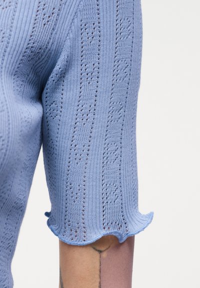 Ανοιχτό μπλε πλεκτό κοντομάνικο με μοτίβο οπών και βελονιές με βολάν στο χέρι ενός ατόμου πάνω σε λευκό φόντο.