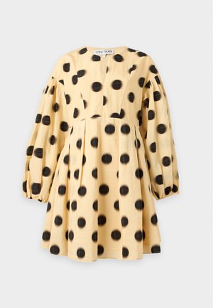 Femme portant une robe jaune ample à gros pois noirs avec de longues manches bouffantes, associée à des ballerines plates noires pointues.