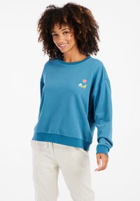 Modrá mikina s okrúhlym výstrihom, dlhými rukávmi a voľným strihom. Na hrudi má farebný grafický logotyp. Mäkká textúra látky.