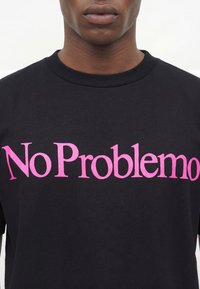 Άνδρας που φοράει μαύρο T-shirt με μεγάλο ροζ κείμενο που λέει «No Problemo» στο στήθος, με λευκό φόντο.