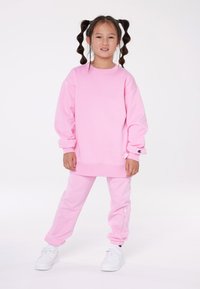 Rozā sporta krekls un atbilstošas bikses, abas izgatavotas no mīkstas auduma. Sporta krekla piedurknes ir nokritušas, bet biksēm ir aproces un logotipa detaļa.