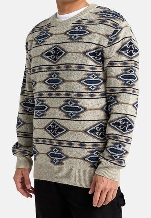 Mand iført beige strikket sweater med blå og brune geometriske mønstre, kombineret med sorte bukser, mod ensfarvet baggrund.