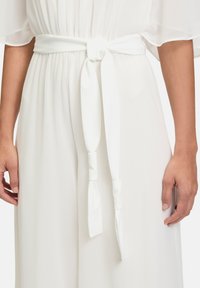 Fehér, folyékony anyagú ruha, megkötött derékövvel. Puha tapintás; hosszú, laza ujjak. Minimális dizájn, amely az elegáns egyszerűségre helyezi a hangsúlyt.