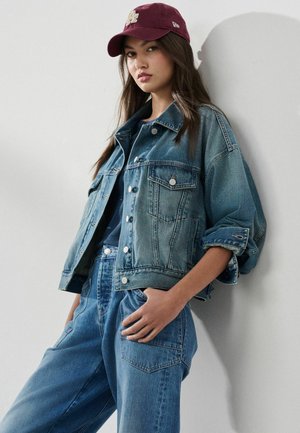 Junge Frau mit einer burgunderroten Mütze, einer übergroßen Jeansjacke mit hochgekrempelten Ärmeln und locker sitzenden Jeans, die an einer weißen Wand lehnt.