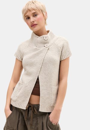 Jonge vrouw met kort blond haar draagt een beige gebreide mouwloze cardigan met twee knopen en zakken, gecombineerd met olijfgroene broek.