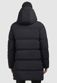 Lang sort puffjakke med hætte, ribbet tekstur, quiltet design og to sidelommer med lynlås. Ærmerne er tætsiddende og slutter over håndleddet.