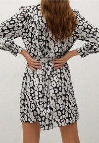 Mujer con cabello largo que lleva un vestido de flores en blanco y negro, con las manos apoyadas en la parte baja de la espalda, de pie frente a un fondo plain.