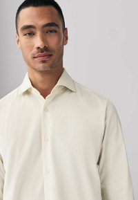 Chemise boutonnée beige clair avec un col pointu et une texture subtile, présentant un plastron avant soigné et des manches longues.