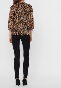 Blouse noire à motifs avec un design floral marron et beige, manches 3/4, associée à un jean skinny noir et des talons noirs.