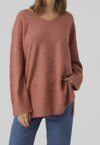Sweter typu V-neck w oversize'owym kroju, wykonany z miękkiego, teksturowanego, czerwono-różowego dzianiny. Posiada opuszczone ramiona i boczne rozcięcia, zestawiony z niebieskimi dżinsami.