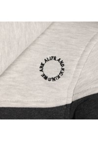 Tela de sudadera en tonos gris y gris claro con un logotipo circular negro bordado. Superficie texturizada con un diseño de bloques de color.