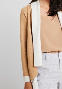 Strickjacke mit einem warmen, sandfarbenen Körper und einem hellgrauen Schalkragen, mit einem lockeren Schnitt und betonten Bündchen. Kombiniert mit einem beigen Oberteil.
