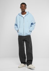 Hellblaues Zip-Hoodie mit entspannter Passform, großen Fronttaschen und Kapuze mit Kordelzug, kombiniert mit schwarzen weiten Hosen und weißen Turnschuhen.