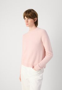 Un pull rose clair avec un col rond, des manches longues, des poignets et un ourlet côtelés, fabriqué à partir d'un tissu doux et texturé. Porté par-dessus un pantalon blanc.