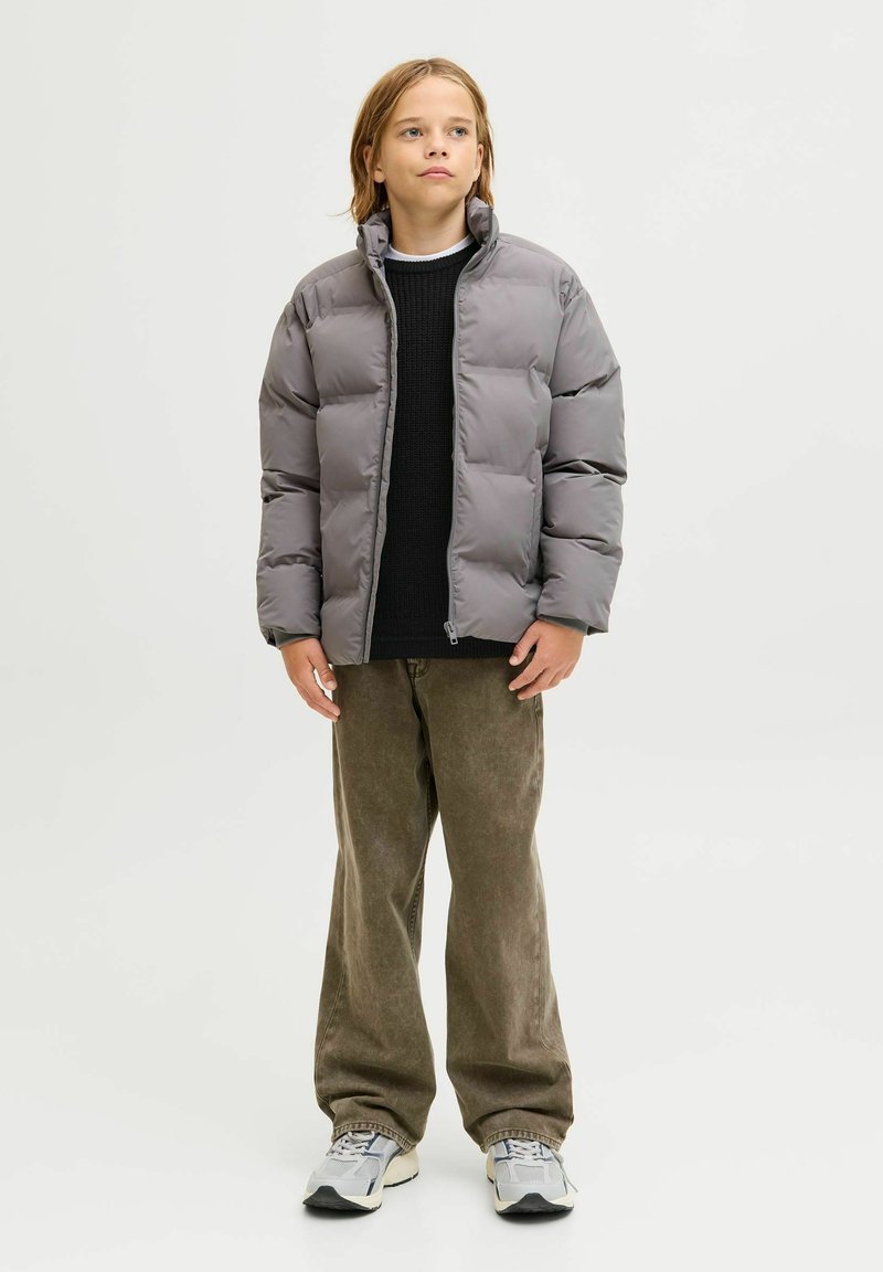 Pufferjacke in sanftem Grau mit hohem Kragen, getragen über einem schwarzen Strickpullover; kombiniert mit olivgrünen, weit geschnittenen Hosen und grauen Sneakers.