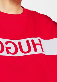 Sudadera roja con cuello redondo que presenta una banda horizontal blanca destacada que muestra la palabra "HUGO" en letras rojas. Textura de tela suave.