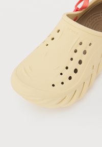 Lys gul Crocs-sko med tekstureret overflade, store ventilationshuller og et formet design. Har en rød hælrem for sikker pasform.