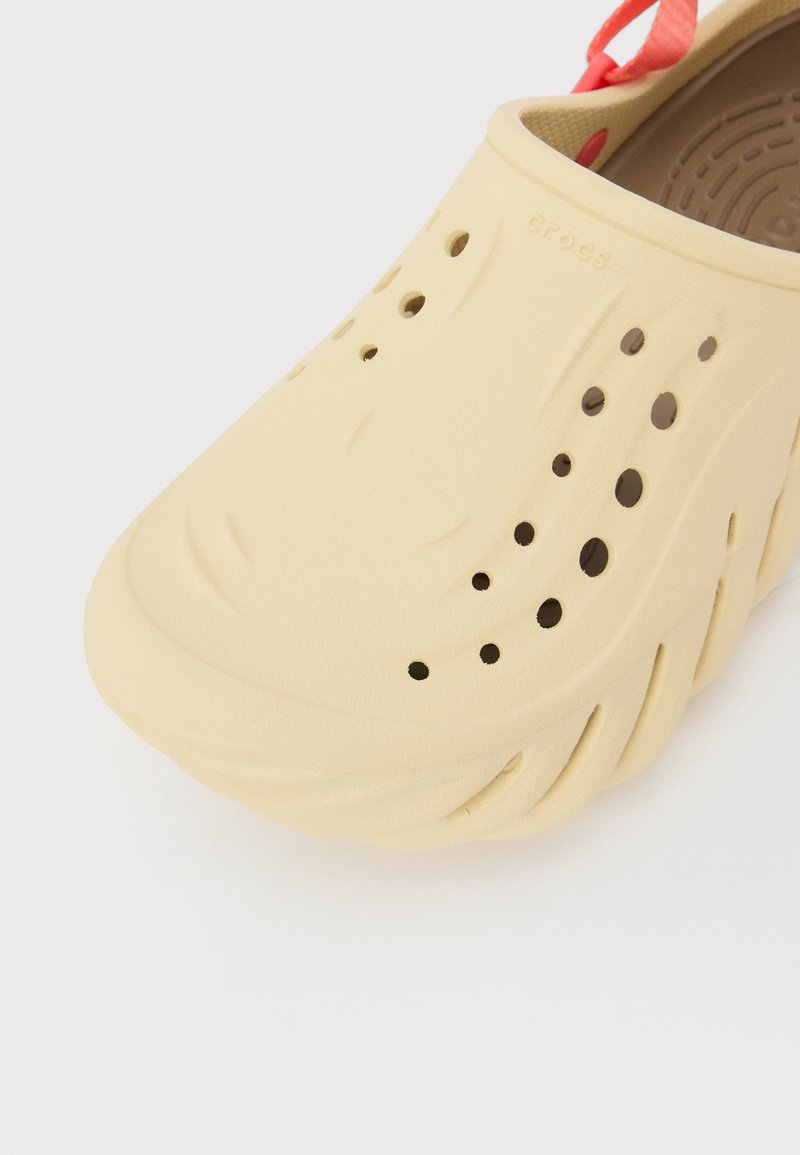 Zapatilla Crocs de color amarillo claro con superficie texturizada, grandes agujeros de ventilación y un diseño moldeado. Presenta una correa en el talón roja para un ajuste seguro.