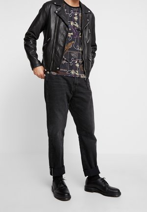 Man in een zwarte leren jas, met een patroon op zijn shirt, opgerolde zwarte jeans, zwarte sokken en zwarte lederen schoenen, staande tegen een witte achtergrond.