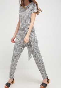 Jumpsuit a motivi bianco e nero con scollatura ampia, maniche corte e una cintura annodata in vita, abbinato a scarpe aperte nere.