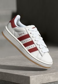 Deportivas blancas con rayas rojas, parte superior de cuero, forro textil, suela de goma y perforaciones. Presenta cordones y un logotipo en el lateral.