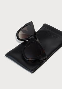 Gafas de sol negras de gran tamaño con un patrón de carey, reposando sobre un estuche de cuero negro grabado con "QUAY". Textura suave, forma angular.