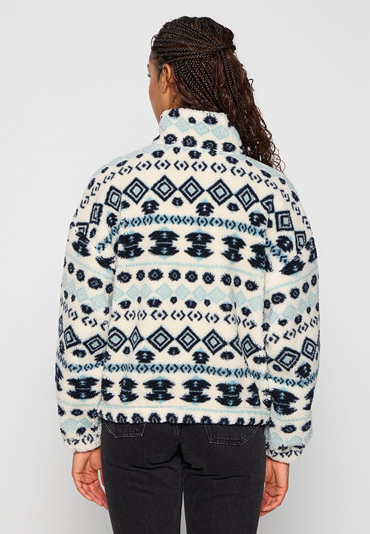 Pull en polaire avec un col haut, présentant un motif géométrique bleu et noir sur fond crème. Texture douce et coupe décontractée.