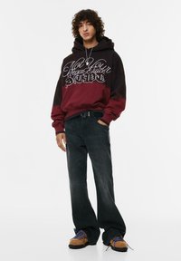 Mikina s kapucňou v čiernej a bordovej farbe, s bielym grafickým textom. Kombinovaná s čiernymi širokými džínami a hnedými teniskami s modrými šnúrkami.