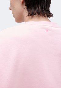 Άτομο με κοντά βρεγμένα μαλλιά που κοιτάζει προς τα πίσω, φοράει ένα ανοιχτό ροζ πουλόβερ με μικρό κεντητό λογότυπο κοντά στο λαιμό.