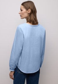 Chemise bleu clair à manches longues avec un col rond, texture douce, et ourlet arrondi, associée à un jean bleu foncé. Design minimaliste.