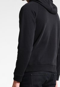 Černá mikina s kapucí z měkké tkaniny, vybavená stahovací kapucí, žebrovanými manžetami a lemem a volnějším střihem. Pohled zezadu zdůrazňuje čisté linie.