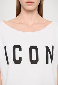 T-shirt blanc surdimensionné avec un décolleté décalé, arborant un texte noir audacieux "ICON" dans une police géométrique. Tissu en coton doux.