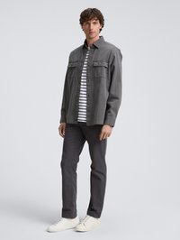 Homme portant une veste grise à boutons sur une chemise rayée noire et blanche, un pantalon gris foncé et des baskets blanches, se tenant contre un fond uni.