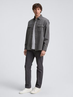 Homme portant une veste grise à boutons sur une chemise rayée noire et blanche, un pantalon gris foncé et des baskets blanches, se tenant contre un fond uni.