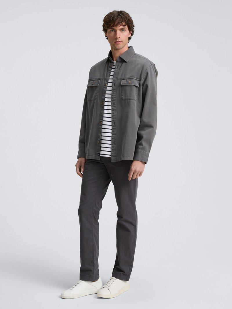 Homme portant une veste grise à boutons sur une chemise rayée noire et blanche, un pantalon gris foncé et des baskets blanches, se tenant contre un fond uni.