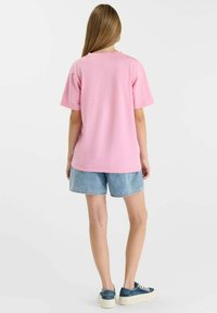Jeune personne debout, portant un t-shirt à manches courtes rose uni, un short en jean bleu clair et des baskets bleu marine avec des semelles blanches.