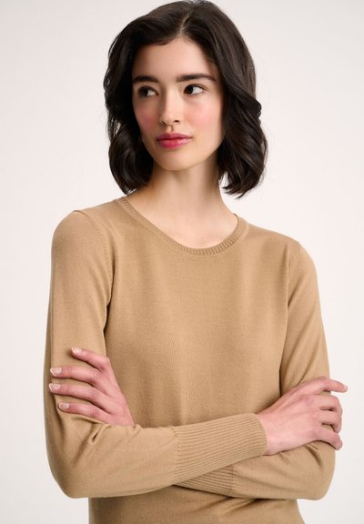 Svetlo rjav pulover z okroglim izrezom, dolgimi rokavi, rebrastimi manšetami in mehko, elastično strukturo. Model pozira z crossed arms.