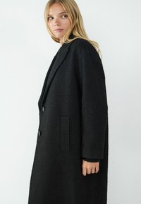 Zwarte jas van een textuurstof; heeft een lange, losse fit, notch-lapels, twee knopen en zijzakken.