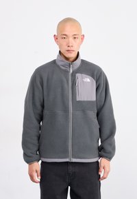 Uomo che indossa una giacca a zip grigia in poliestere con tasca sul petto e pantaloni neri, in piedi su uno sfondo bianco semplice.