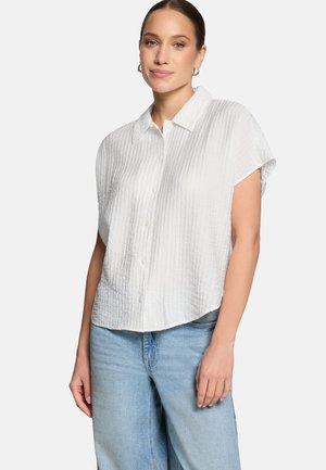 Femme aux cheveux attachés porte une chemise blanche texturée à manches courtes et un jean bleu clair, debout devant un fond uni.
