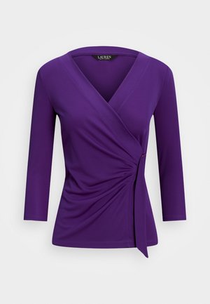 Fioletowa bluzka z dekoltem w kształcie litery V i rękawami o długości trzy czwarte. Posiada miękki, gładki materiał oraz marszczenie po lewej stronie z wiązaniem.